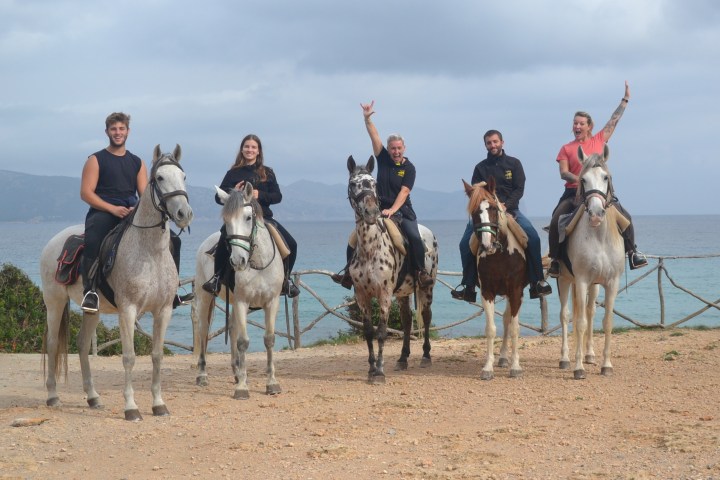 Five people on horses by the sea with cloudy sky, some raising arms in celebration.