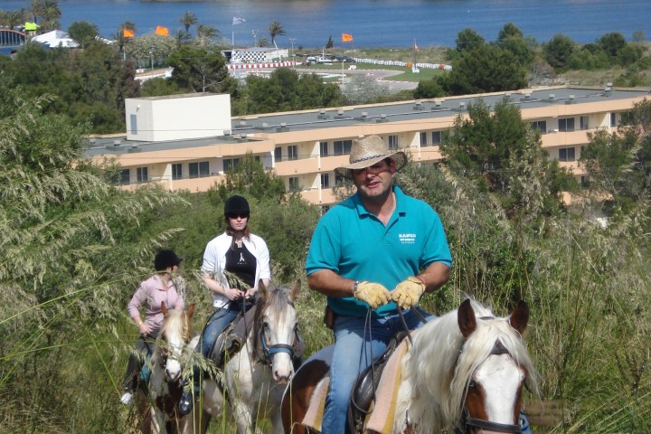 Three people riding horses on a grassy hillside with a town and water in the background.