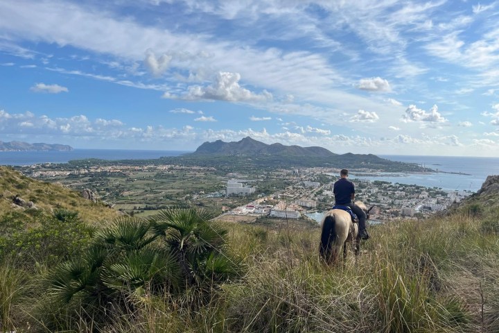 Person on horseback overlooking a coastal town with mountains and sea in the background.
