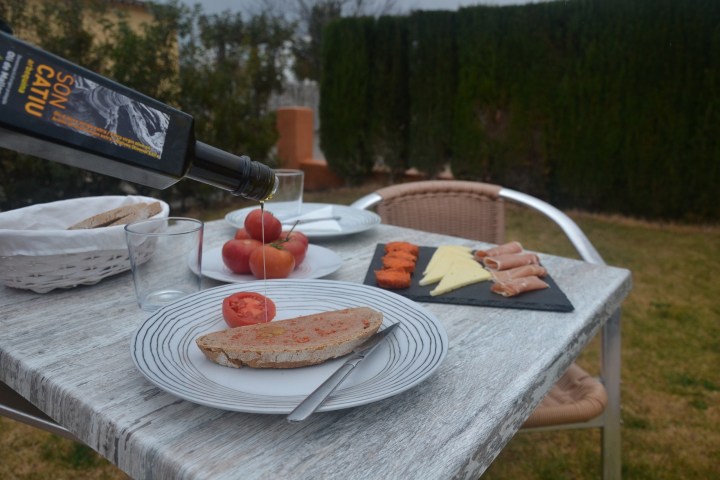Outdoor table with bread, tomatoes, cold cuts, cheese, and olive oil being poured on bread.