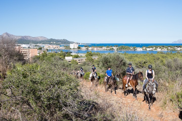 Group of people riding horses on a trail with a coastal city and ocean in the background.