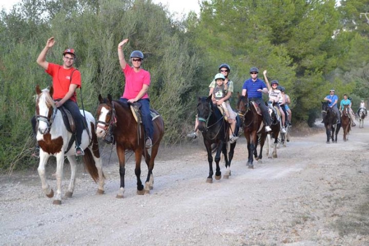 Group of people riding horses on a dirt path, waving, with trees in the background.